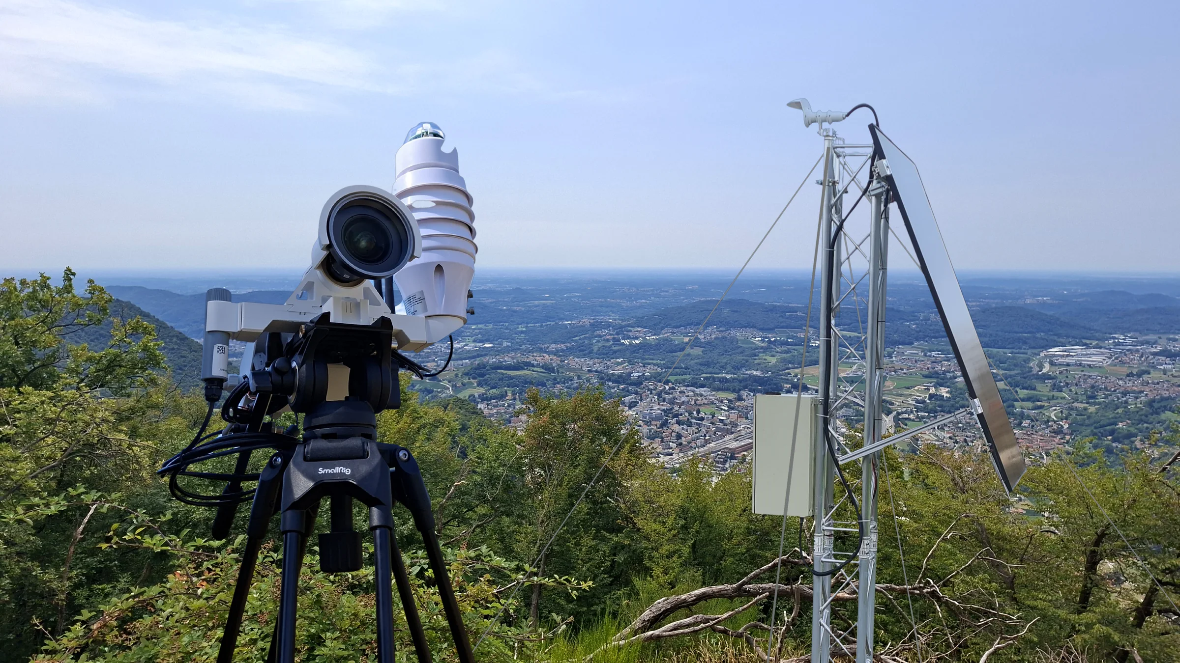 HUVI.NOMAD auf einem Stativ im Vordergrund, dahinter ein HUVI.GUARDIAN Sensorturm mit Kamera und Solarpanel, im Hintergrund das Tessin